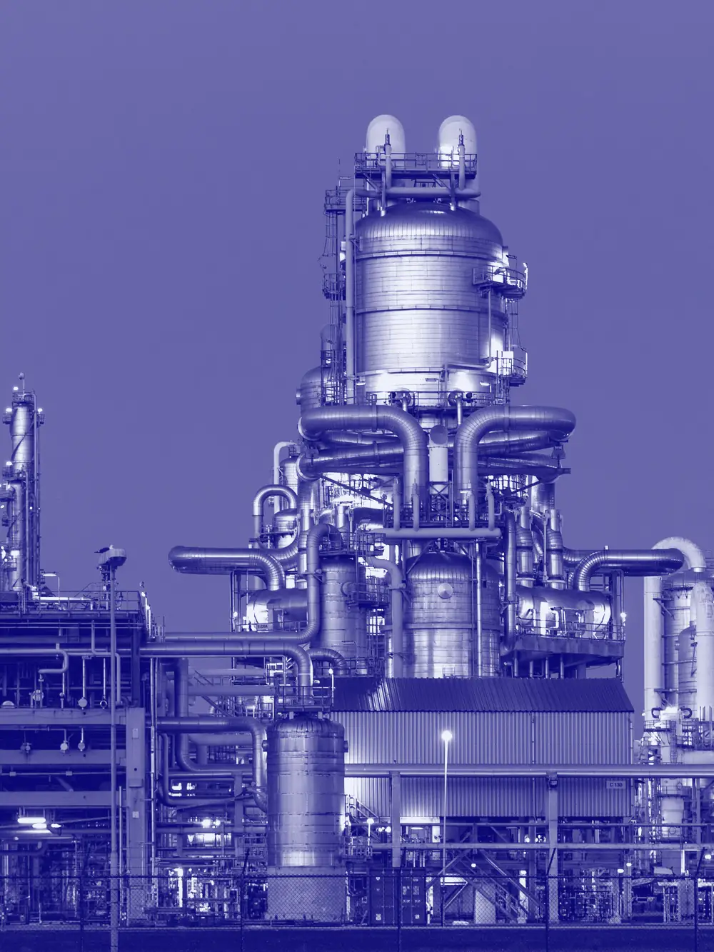 A process plant at dusk, stacks and pipework lit against the sky